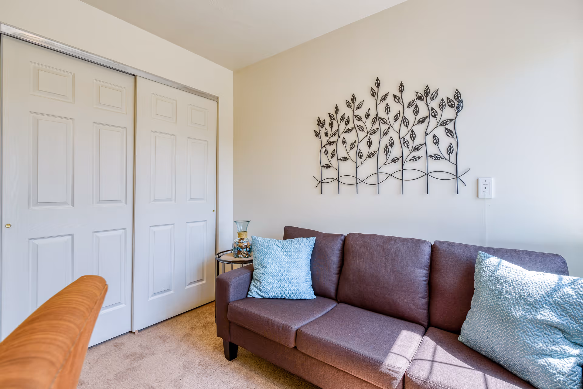 A cozy living room area featuring a brown sofa with two light blue textured pillows. Above the sofa is a decorative metal wall art piece with a leaf design. To the left of the sofa is a small round side table with a glass vase containing decorative items. The background shows white sliding closet doors and beige carpeted flooring.