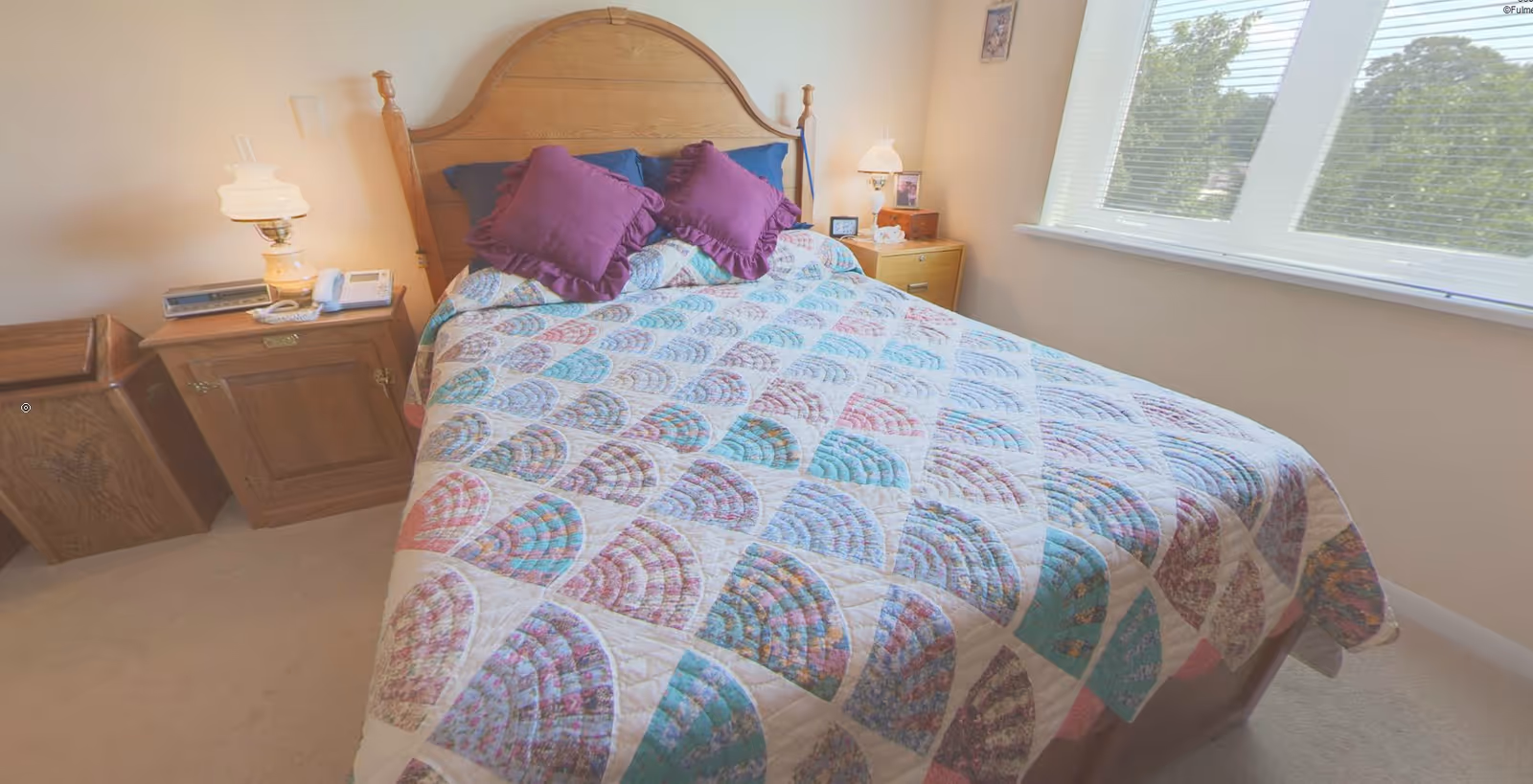 A bright bedroom with a wooden headboard bed covered by a patterned quilt and purple pillows, flanked by nightstands and a window.