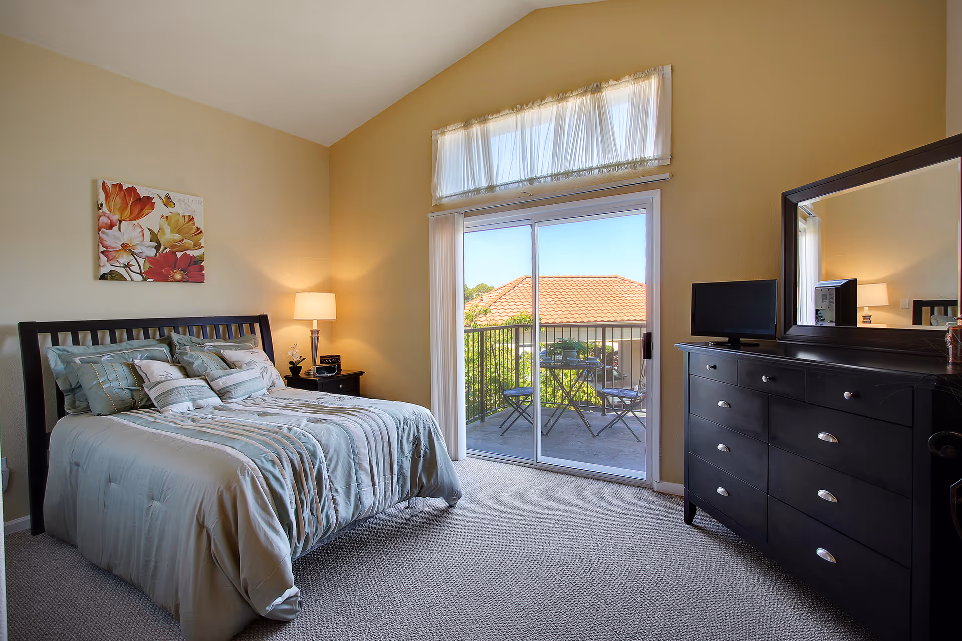 A bedroom with a bed covered in light blue and beige bedding, a nightstand with a lamp and small plant, a black dresser with a mirror and a small TV on top, and a sliding glass door leading to a balcony with a small table and two chairs. The walls are painted beige and there is a floral painting above the bed.
