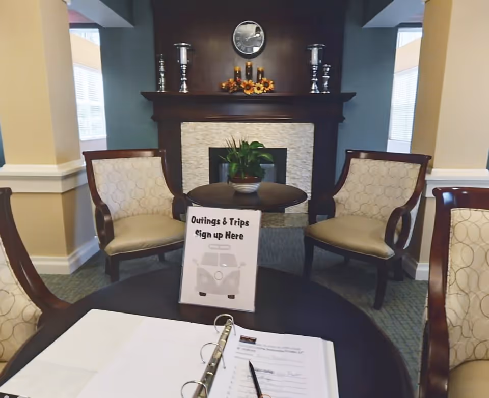 A cozy seating area with four upholstered chairs around a dark wooden table. On the table is a sign that reads 'Outings & Trips Sign up Here' with a picture of a van, and an open binder with a pen. In the background, there is a fireplace with decorative candles and a clock above it, along with a small potted plant on a round table in front of the fireplace.