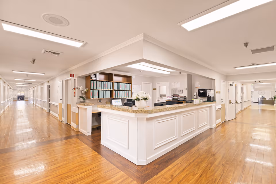 A bright and clean nursing station area in a senior living facility with a long hallway extending on both sides. The nursing station has a white counter with a granite top, office supplies, and a vase with white flowers. The floor is wooden, and the walls are painted white with handrails along the hallway.
