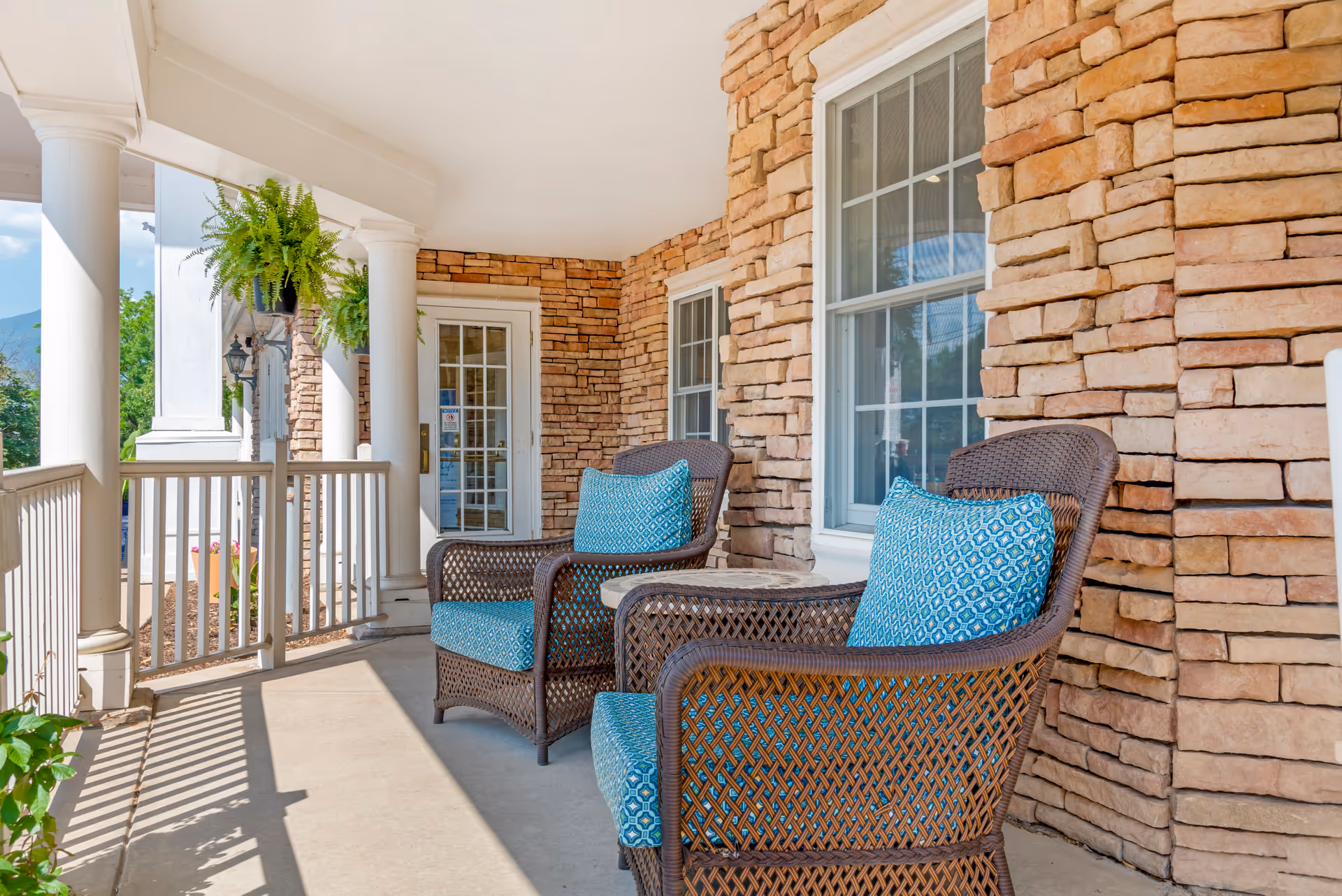 Covered front porch with wicker chairs featuring blue patterned cushions against a stone facade.