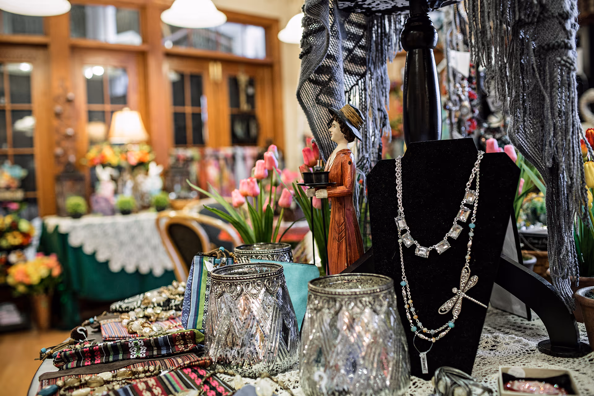 A display table inside a room featuring various decorative items including jewelry on a black necklace stand, a wooden figurine of a woman holding a tray, glass candle holders, and colorful fabric pieces. In the background, there are pink tulip flowers, a table covered with a green and white lace tablecloth, chairs, and wooden cabinets with glass doors.