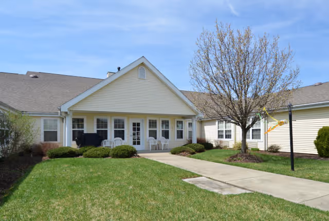 Front of a single-story senior living building with a porch, white chairs, a walkway, lawn, and a tree.