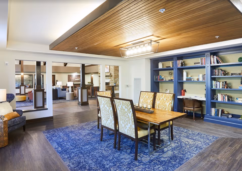 A well-lit interior room with a wooden dining table surrounded by six upholstered chairs with yellow patterned fabric. The floor has a blue patterned rug under the table, and the ceiling features a wooden panel with a modern light fixture. In the background, there is a built-in blue bookshelf filled with books and decorative items, and a small desk with a chair. The room opens into another seating area with various chairs and a stone fireplace.