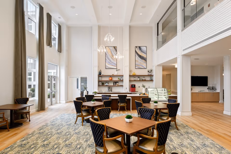 A bright and spacious dining area with high ceilings and large windows. The room features several wooden tables with cushioned chairs arranged around them. There is a counter at the back with shelves holding decorative items and a small display case. Modern pendant lights hang from the ceiling, and the floor is covered with a patterned carpet and light wood flooring.