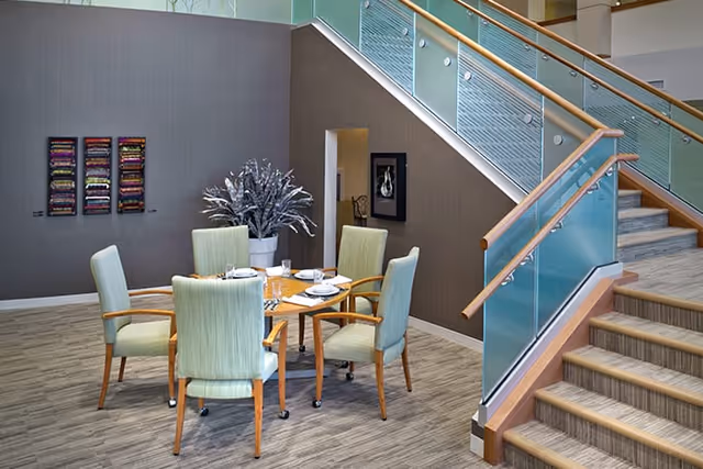 A modern interior space featuring a round wooden table set with plates, glasses, and napkins, surrounded by six light green upholstered chairs with wooden arms. To the right, there is a staircase with glass panels and wooden handrails. On the left wall, there are three colorful framed artworks and a large potted plant with silver leaves.