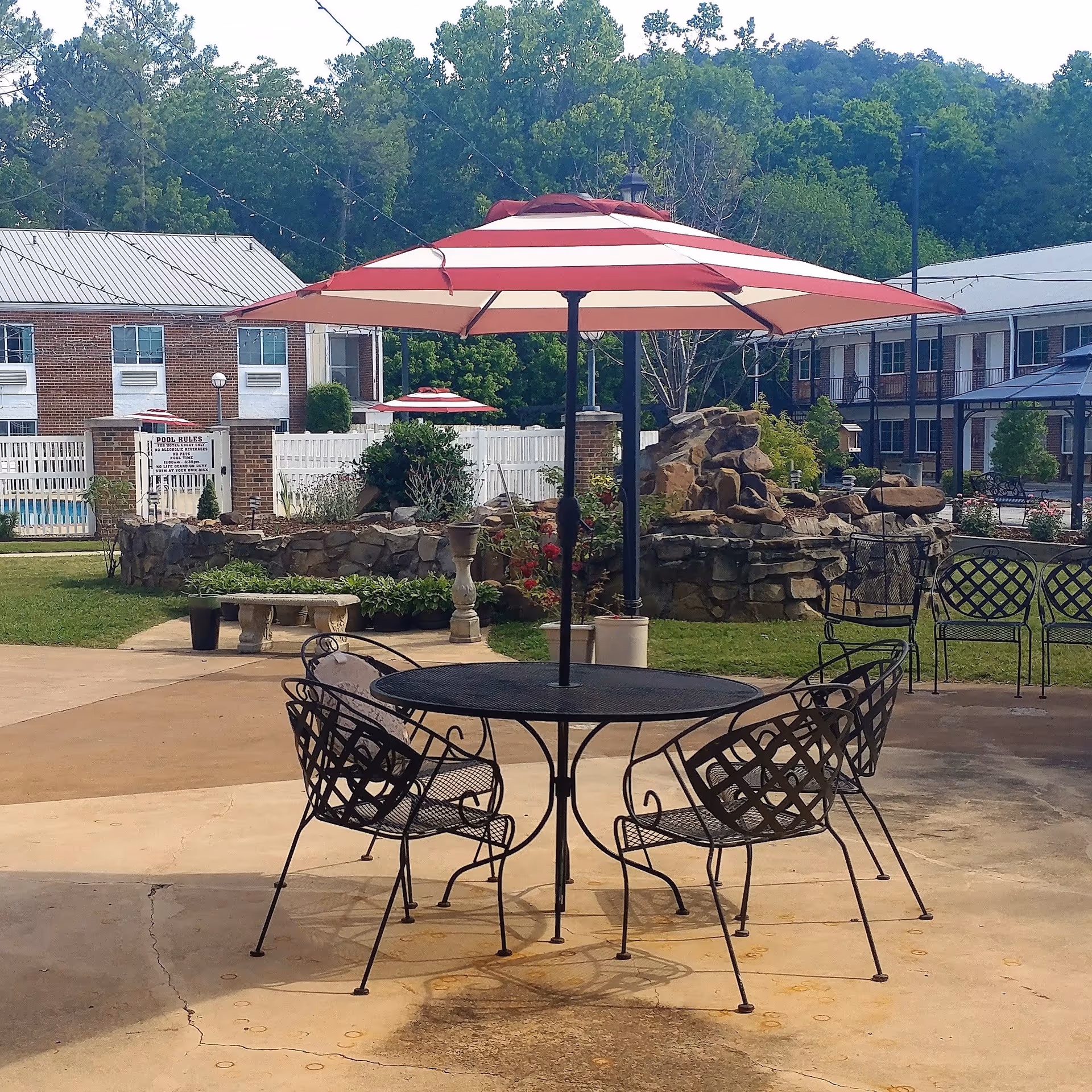 Outdoor patio area with a round metal table and four matching chairs under a large red and white striped umbrella. In the background, there is a rock water feature, greenery, and two brick buildings with white fences and windows. String lights are hung above the patio area.