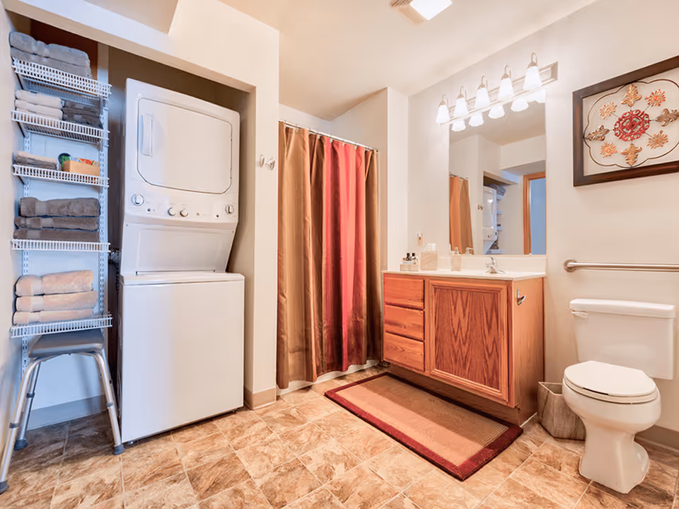 A bathroom with a stacked washer and dryer unit on the left, a metal shelving unit holding folded towels, a shower with a brown and red curtain, a wooden vanity with a sink and mirror above it, a toilet with a grab bar beside it, and a framed decorative artwork on the wall.