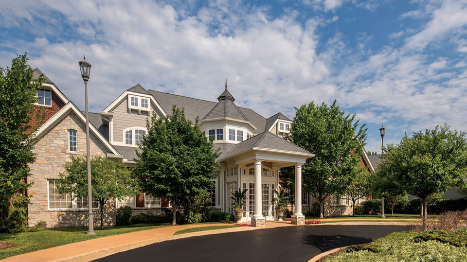 Front entrance of Parc Provence senior living building with a porte-cochère, trees, and a curved driveway under a partly cloudy sky.