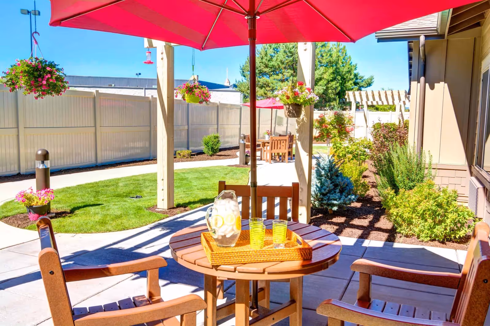 Sunny outdoor patio with a round wooden table and chairs under a red umbrella, a pitcher and glasses on the table, hanging flower baskets, and landscaped paths.