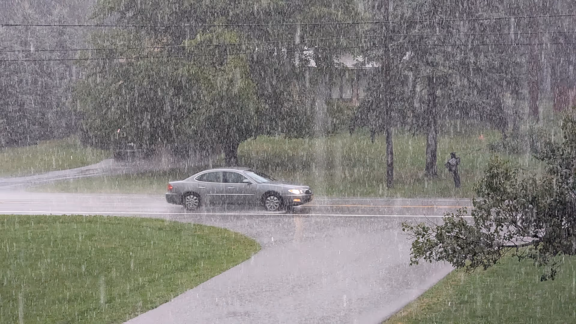 A silver sedan drives along a wet road in heavy rain with trees, grass, a driveway in the foreground, and a person standing on the sidewalk.