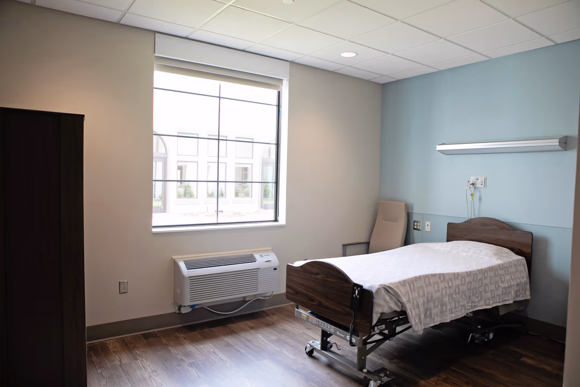 A simple, clean bedroom in a senior living facility with a single hospital-style bed covered with a white patterned blanket, a beige chair in the corner, a large window letting in natural light, and a wall-mounted air conditioning unit below the window. The walls are painted light blue and beige, and the floor has wood-like flooring.