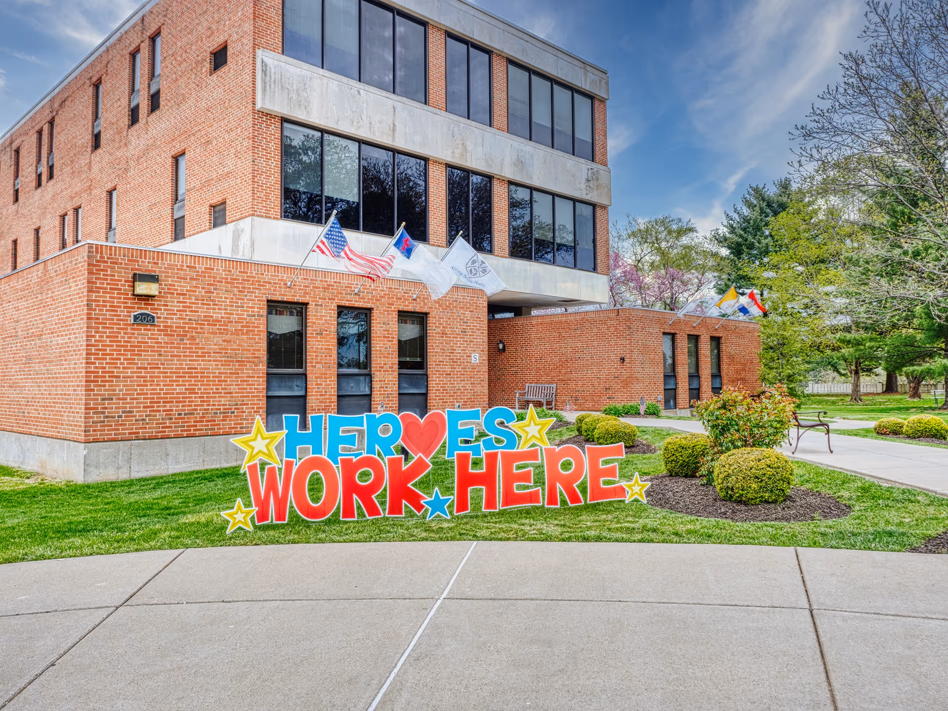 Brick multi-story senior facility building with flags and a large colorful "HEROES WORK HERE" sign on the front lawn.