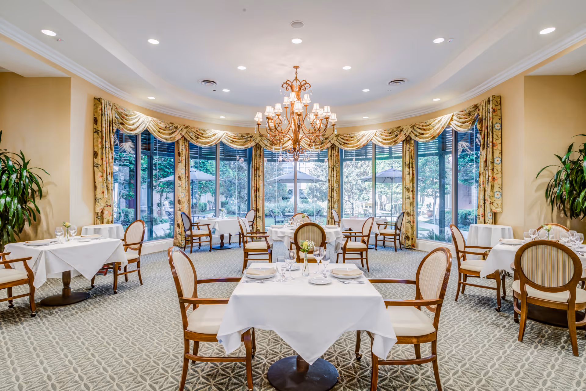 Elegant dining room with round tables covered in white tablecloths, set with plates, glasses, and silverware. The room features large windows with floral curtains overlooking a garden with umbrellas, a patterned carpet, and a central chandelier hanging from the ceiling.