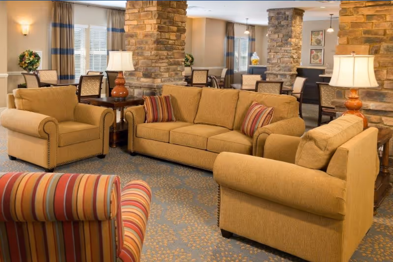 A cozy living room area with mustard yellow sofas and armchairs, including a striped armchair in the foreground. The room features stone pillars, side tables with lamps, and a carpet with a subtle pattern. In the background, there are dining tables and chairs near windows with curtains.