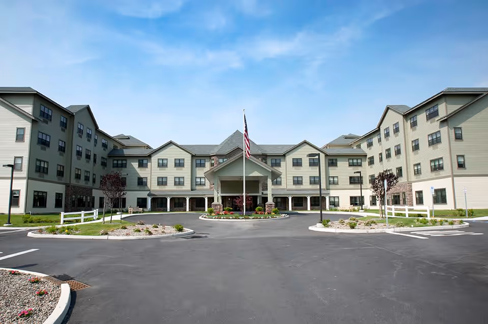 Front exterior view of Braemar at Wallkill, a multi-story senior living facility with a central entrance, landscaped roundabout with an American flag, and parking areas on either side under a blue sky.