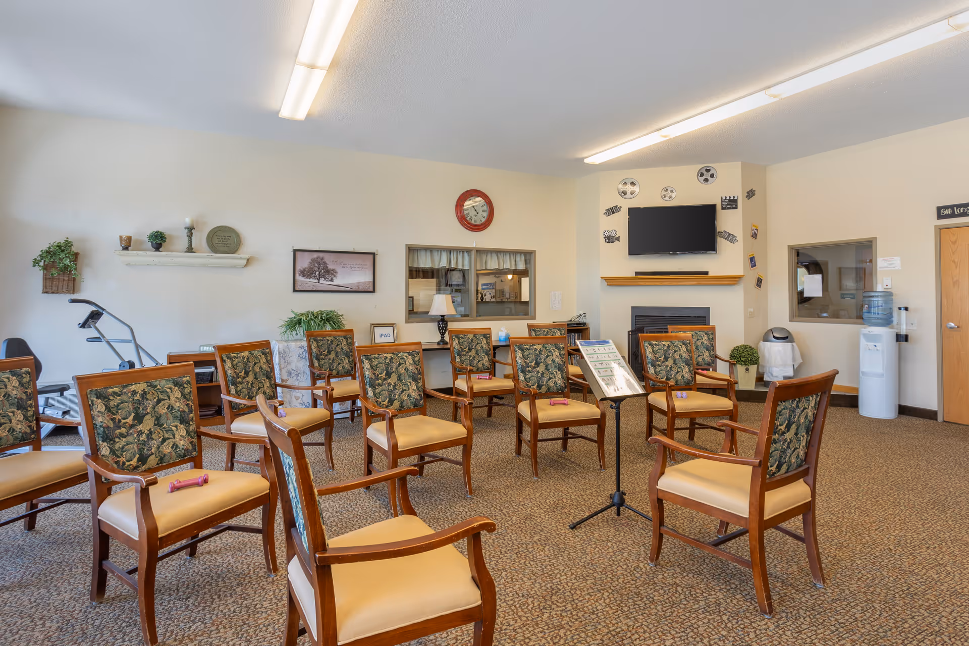 A spacious room with multiple wooden chairs arranged in a semi-circle, each with floral-patterned backs and beige seats. A music stand with sheet music is positioned in the center. The room has beige walls decorated with a clock, framed pictures, and film reel decorations around a mounted flat-screen TV above a fireplace. There is a water cooler and a door on the right side, and exercise equipment is visible on the left side near a shelf with decorative items.