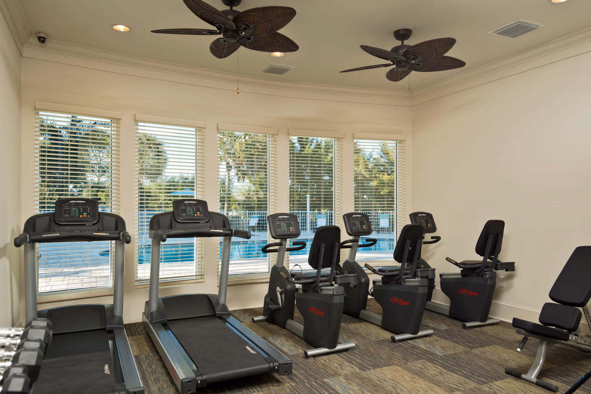 Indoor fitness room with two treadmills, three recumbent exercise bikes, and a weight bench. Large windows with white blinds show an outdoor pool area with palm trees. The room has ceiling fans and neutral-colored walls and carpet.