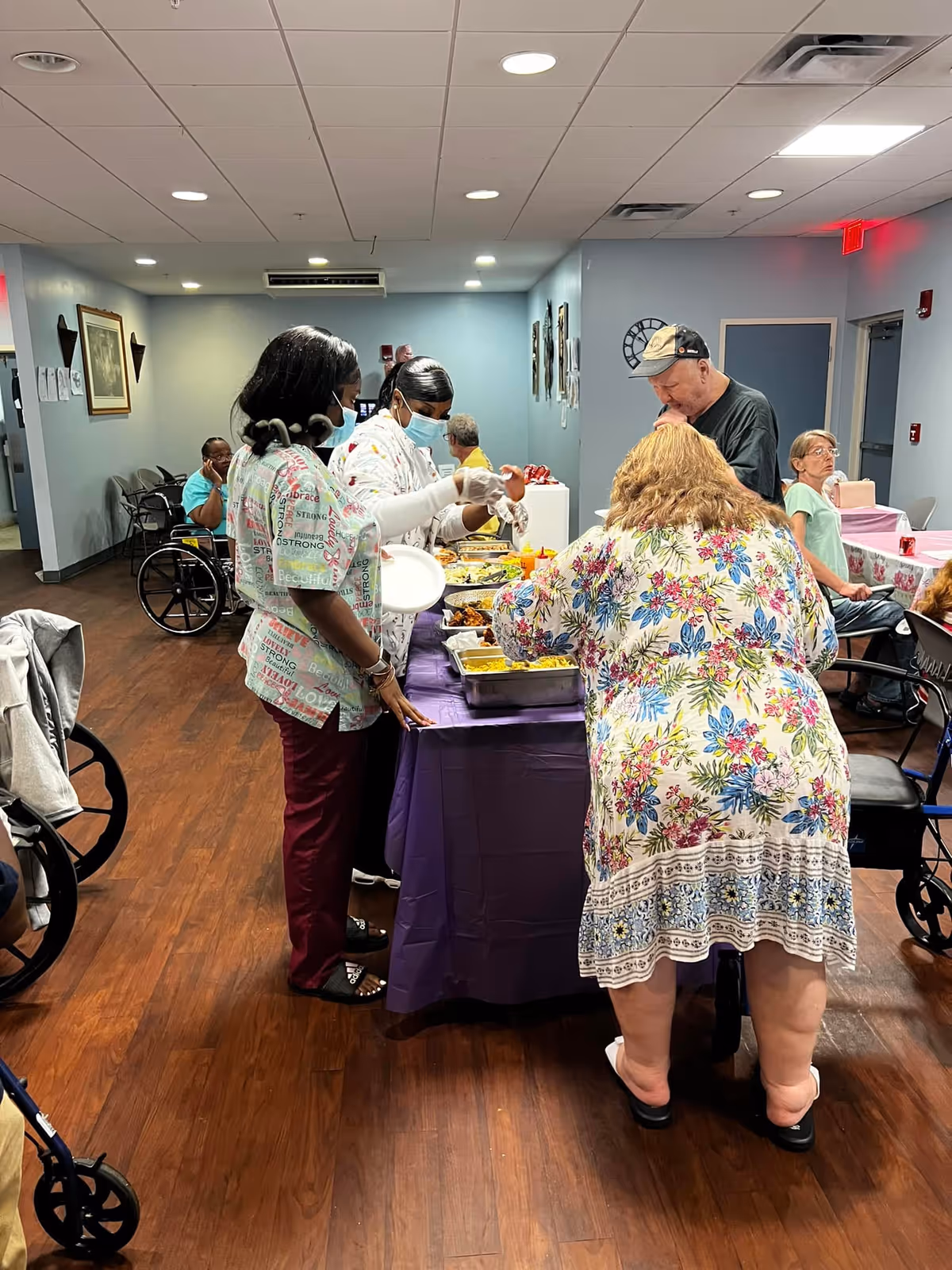 Several people, including staff and residents, are gathered around a buffet table with food trays in a senior living facility dining area. The staff are serving food while some residents wait in line or sit nearby. The room has wood flooring, light blue walls, and ceiling lights.