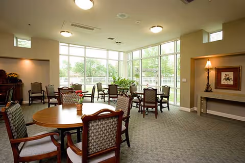 Sunlit communal dining area with round tables, upholstered chairs, and large floor-to-ceiling windows overlooking greenery.