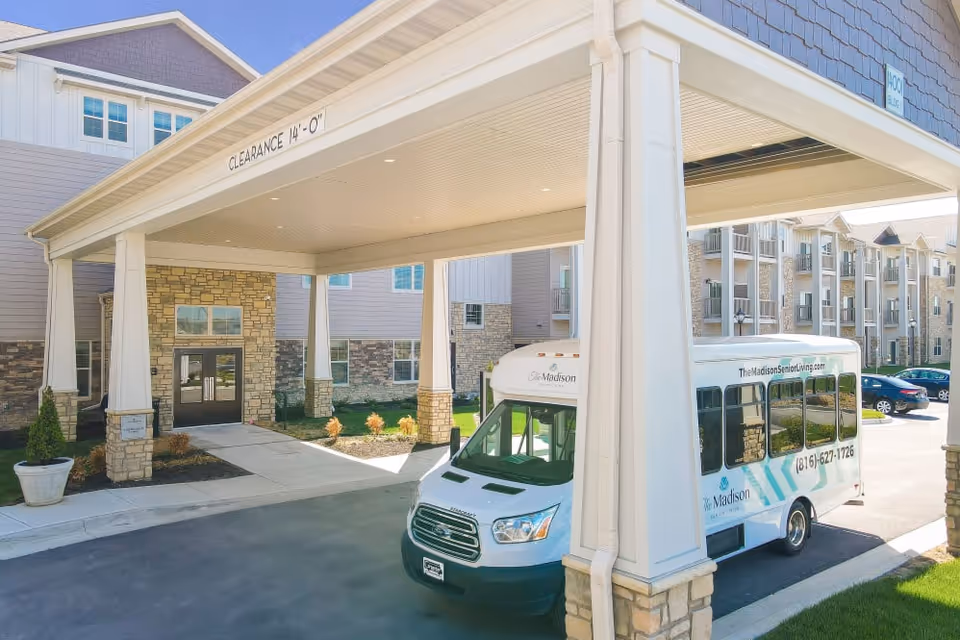 Covered porte-cochère entrance of a senior living building with a small shuttle bus parked underneath and apartment balconies in the background.