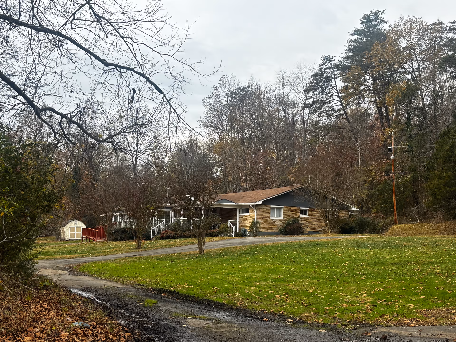 A single-story house with a brown roof and stone facade surrounded by trees with autumn foliage. There is a curved driveway leading up to the house, a small white shed with a red ramp on the left side, and a grassy lawn in the foreground. The sky is overcast.
