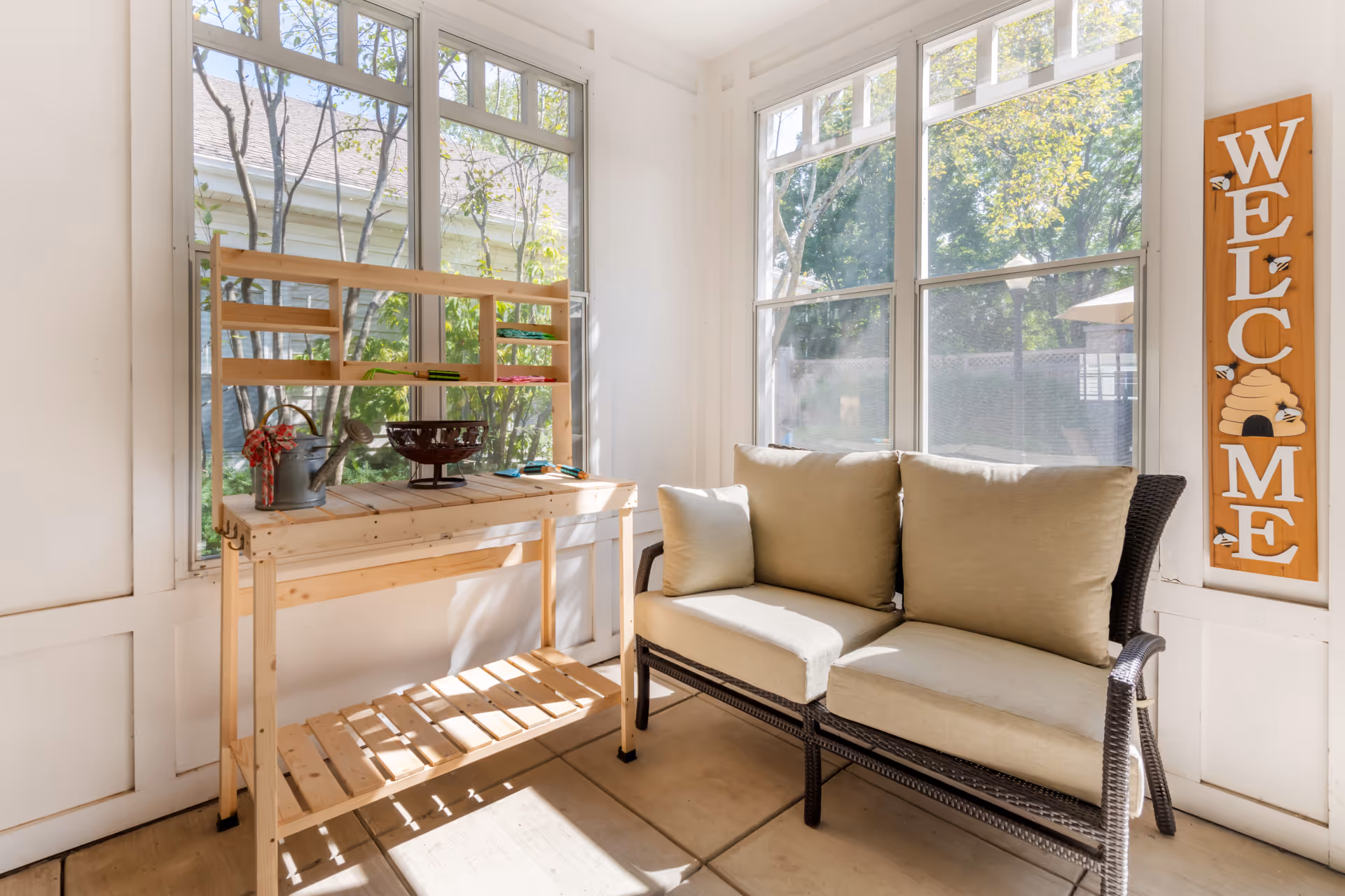 A bright sunroom with large windows showing outdoor greenery. Inside, there is a wooden potting bench with gardening tools and a watering can, and a cushioned wicker loveseat. A wooden sign on the wall reads 'WELCOME' with a beehive and bees decoration.