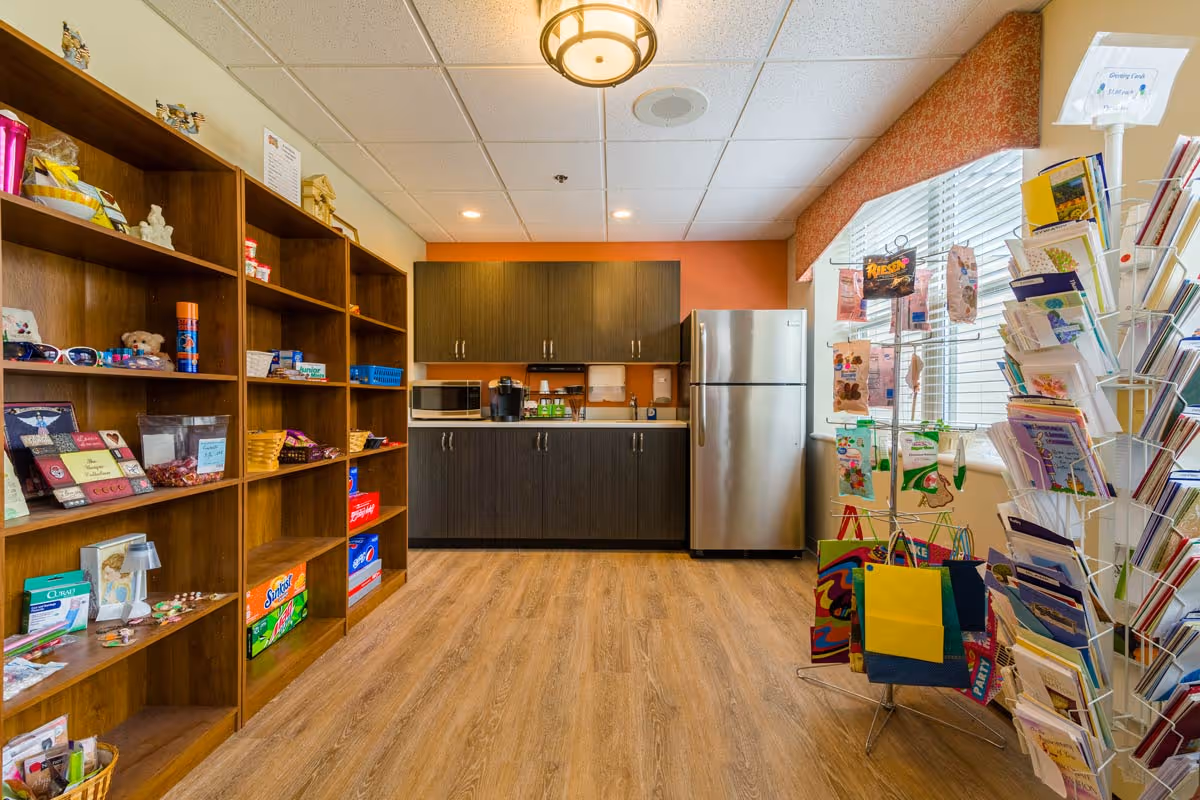 Interior view of a small kitchenette area with wooden shelves on the left holding various items like snacks, sunglasses, and small gifts. In the center back, there are dark wood cabinets, a microwave, a coffee maker, and a stainless steel refrigerator. On the right side, there is a window with blinds and a display rack filled with greeting cards and gift bags on the floor.