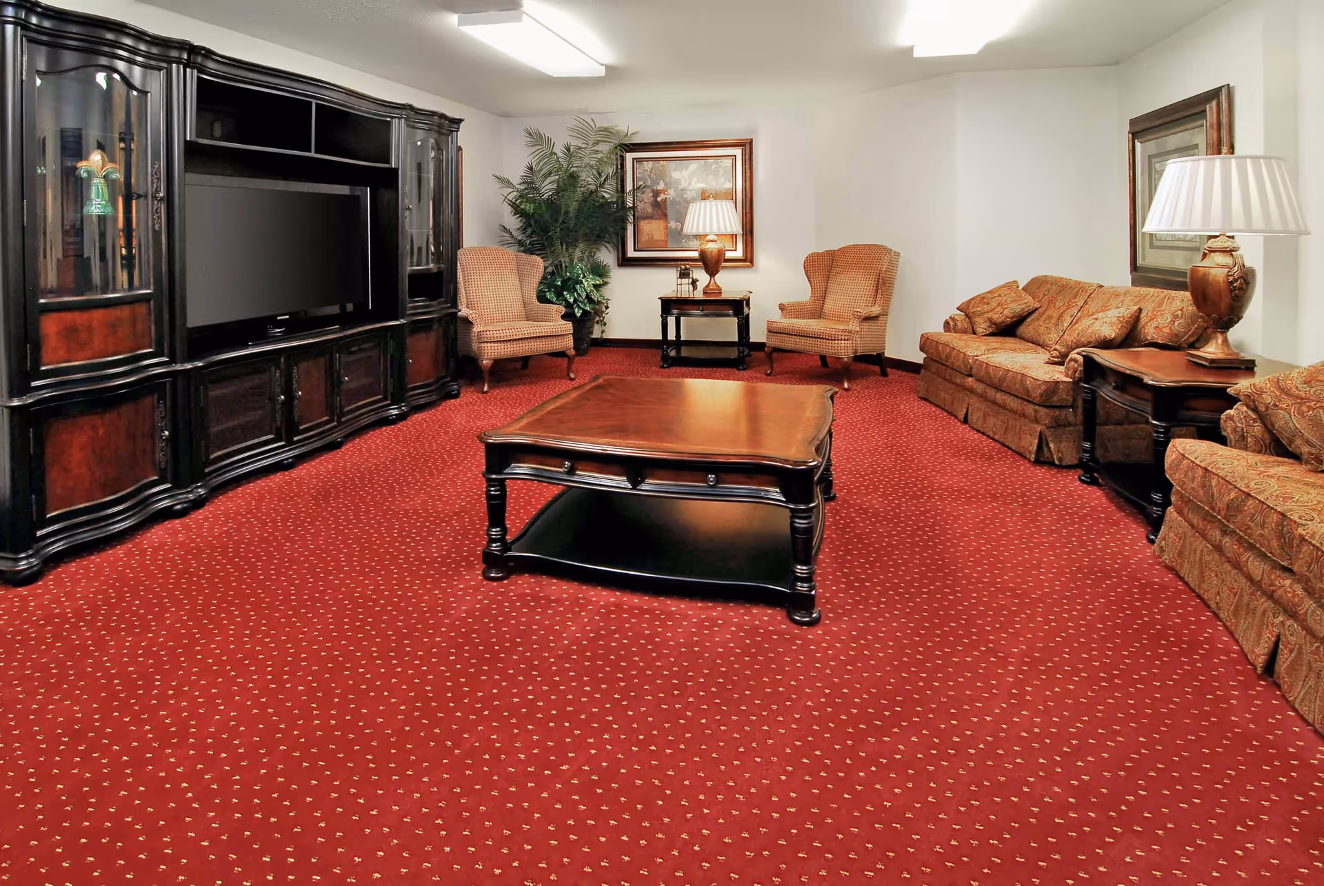 A cozy living room with red patterned carpet, a large wooden entertainment center with a TV, two upholstered armchairs, a wooden coffee table, a sofa with patterned cushions, side tables with lamps, framed artwork on the walls, and a large potted plant in the corner.