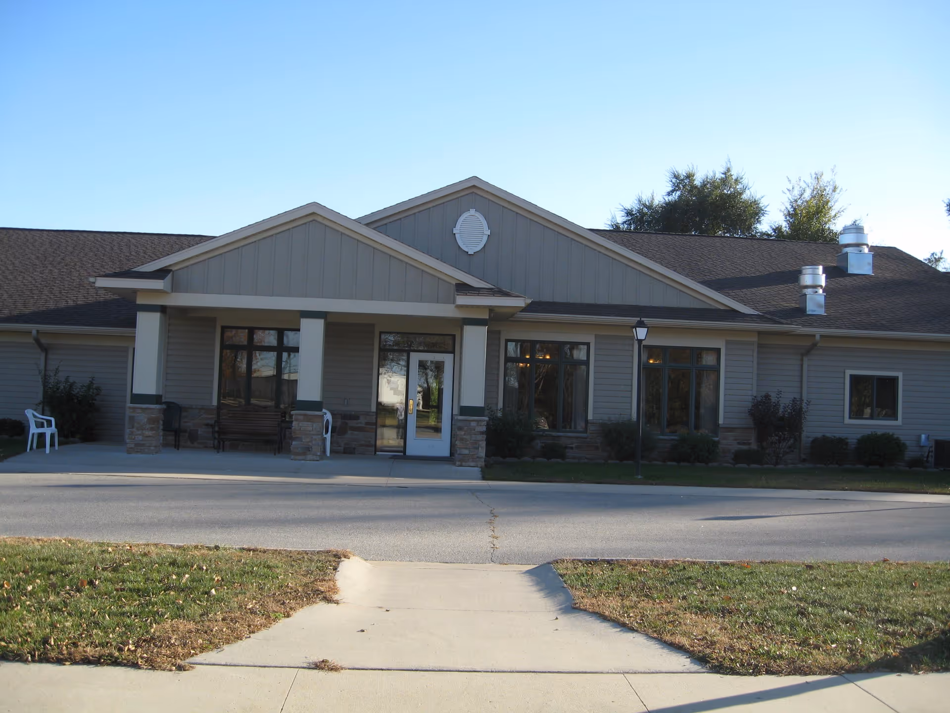 Front exterior view of Cedar Vale Assisted Living facility showing a single-story building with a covered entrance, large windows, and a paved driveway. There are a few chairs and a bench near the entrance, with some shrubs and trees in the background under a clear blue sky.
