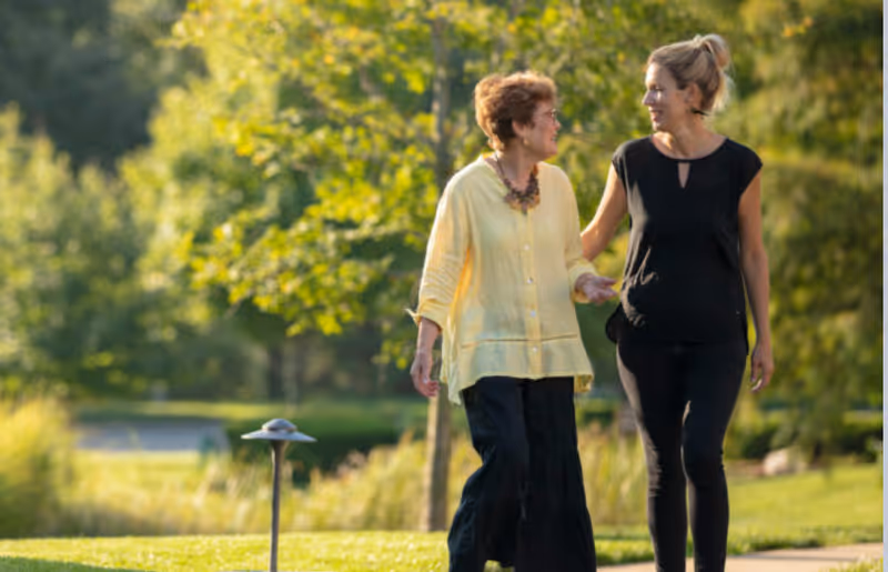 An elderly woman and a younger woman walking together on a paved path in a green outdoor park-like setting, surrounded by trees and grass, engaging in conversation and smiling.