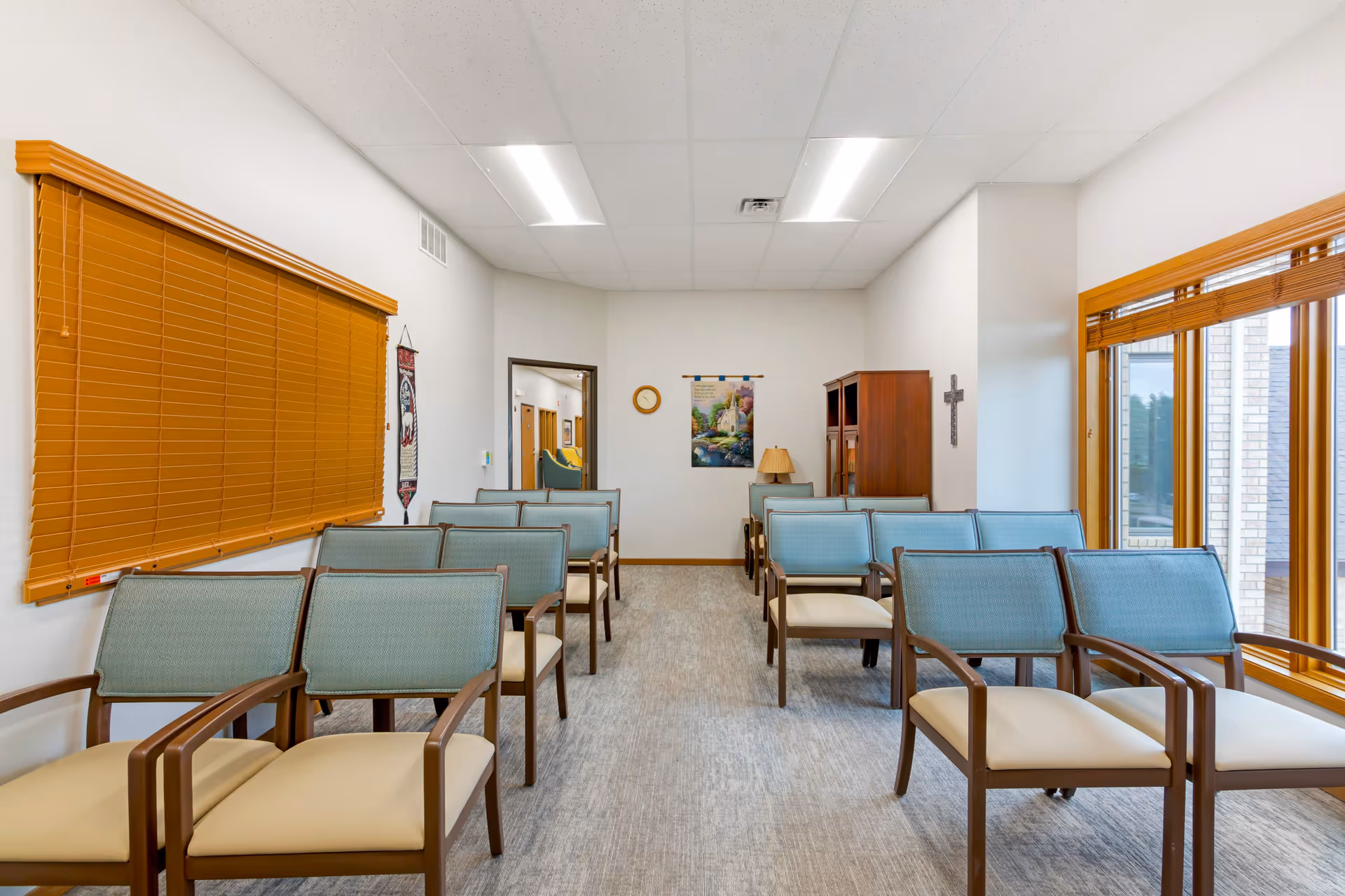 A small chapel or meeting room with rows of wooden chairs with light blue cushions arranged facing a wall with a clock, a framed picture, a lamp on a small table, and a wooden cabinet. The room has white walls, a gray carpeted floor, and large windows with wooden blinds on the right side.