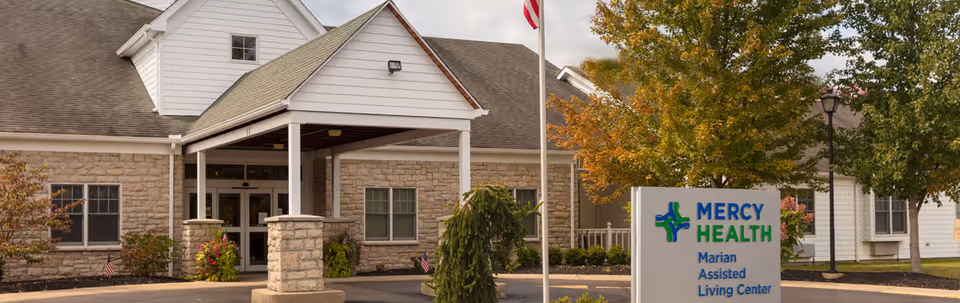 Front entrance of Mercy Health Marian Assisted Living Center with a covered porte-cochere, flagpole, landscaping, and a sign in the foreground.