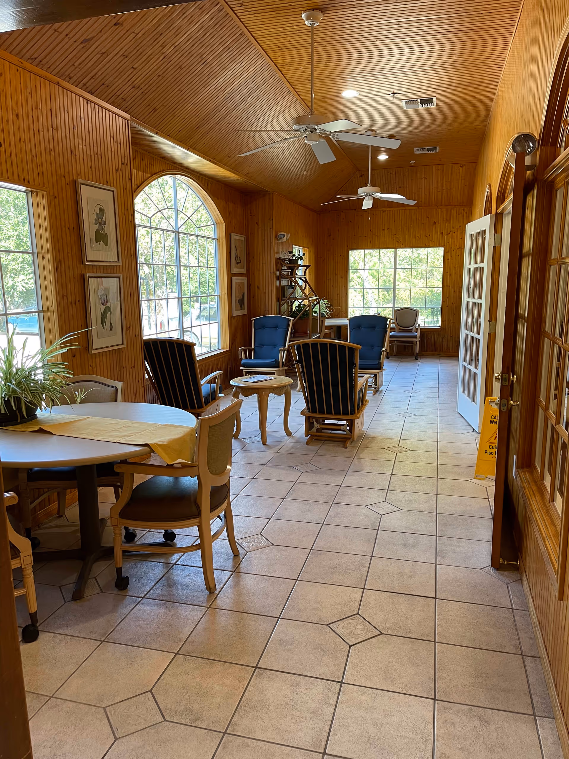 A bright and cozy common area with wooden paneled walls and ceiling, large arched windows letting in natural light, several cushioned chairs arranged around small tables, a round table with chairs in the foreground, ceiling fans, and framed artwork on the walls.