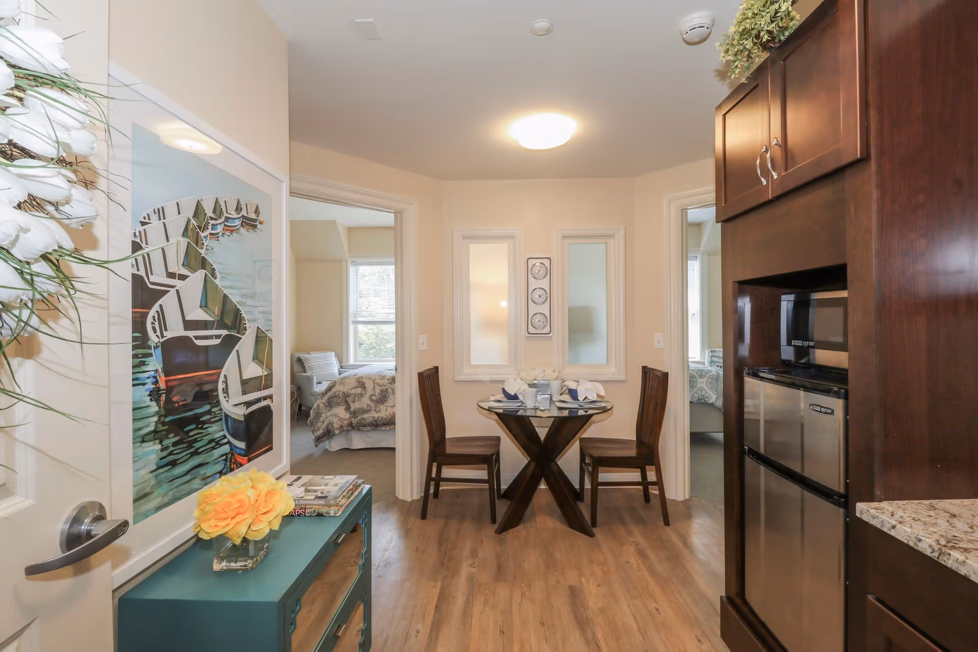 Interior view of a small dining area with a round glass table set for two with napkins and plates. The dining area is flanked by two doorways leading to bedrooms with beds visible. To the right, there is a kitchenette area with a microwave and mini fridge built into dark wood cabinetry. On the left side, a teal cabinet with a vase of yellow flowers and a large framed picture of boats on water is visible. The floor is wooden and the walls are light-colored.