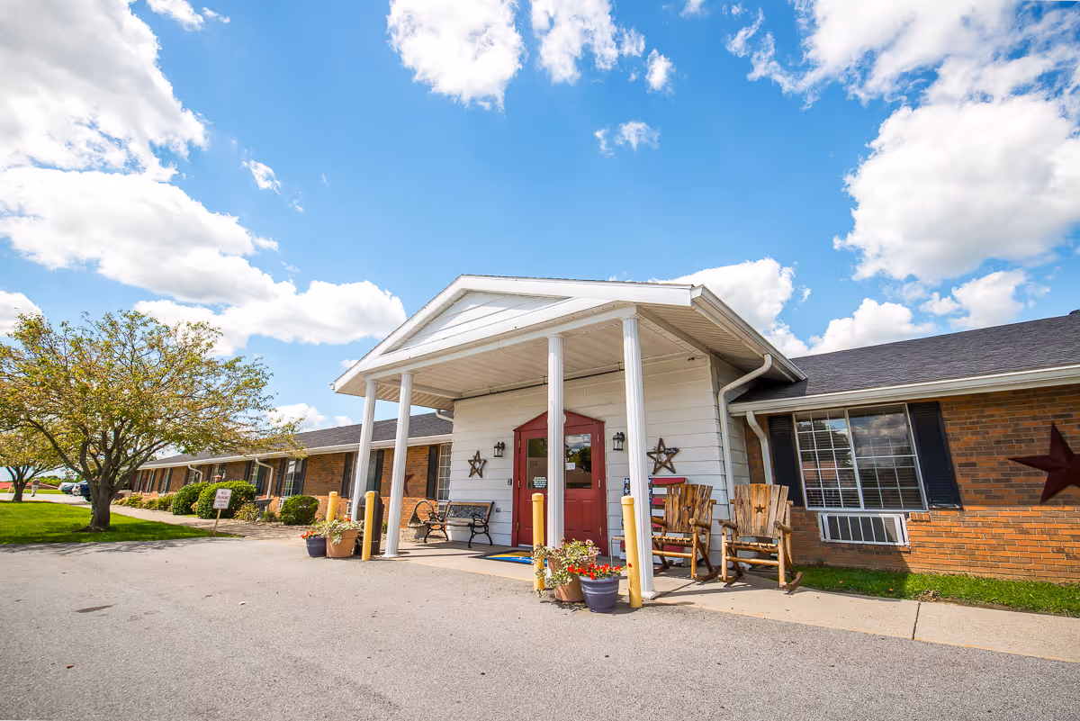 Exterior view of Signature HealthCARE of Trimble County building with a covered entrance supported by white columns, a red door, wooden chairs, potted plants, and a tree on the left side under a partly cloudy blue sky.
