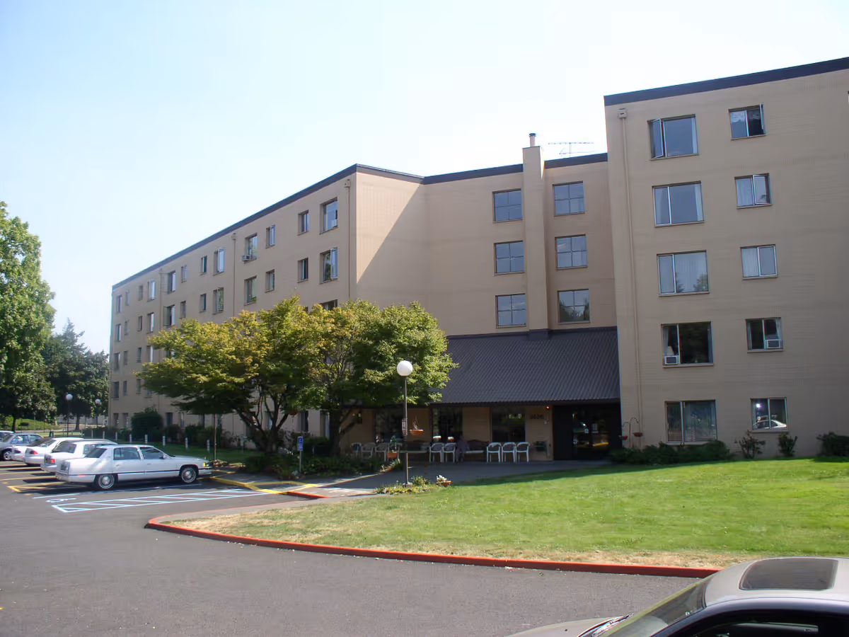Front exterior of a beige multi-story senior living building with a parking lot, lawn, and trees.