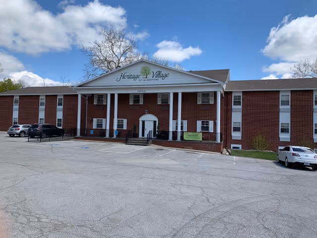 Front exterior view of a two-story brick building with white columns and a sign that reads 'Heritage Village of Gladstone' under a partly cloudy sky. There are several parked cars in the parking lot in front of the building.