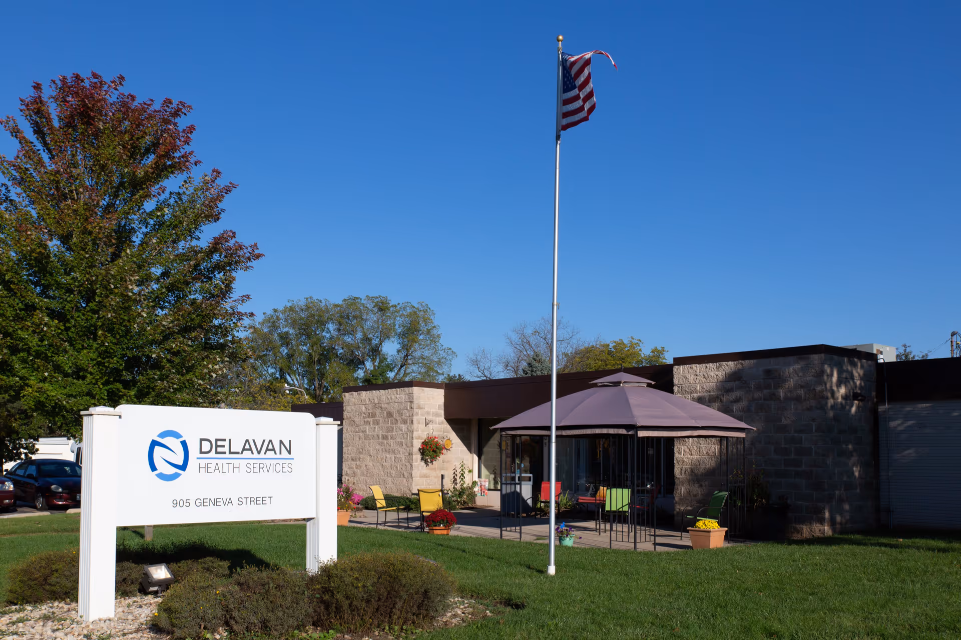 Front entrance of Delavan Health Services building with a sign, flagpole, and outdoor seating under a patio umbrella.