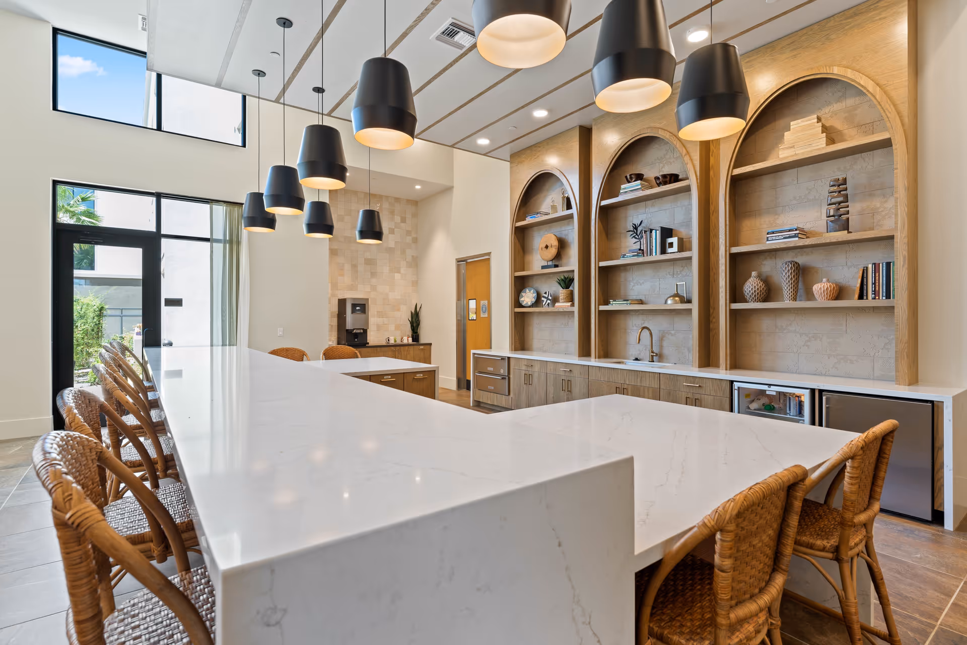 Modern kitchen area with a large white marble island countertop surrounded by wicker chairs. Above the island, there are multiple black pendant lights hanging from the ceiling. The back wall features built-in wooden shelves with decorative items and a small sink. Large windows and a glass door allow natural light to fill the space.