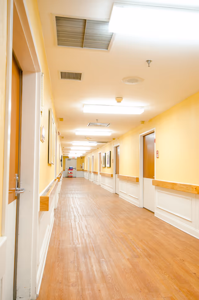A long, well-lit hallway in a senior living facility with wooden floors, yellow walls, white wainscoting, and multiple closed wooden doors on both sides. The ceiling has fluorescent lights and ventilation grilles. There are handrails along the walls and framed pictures hanging on the walls.