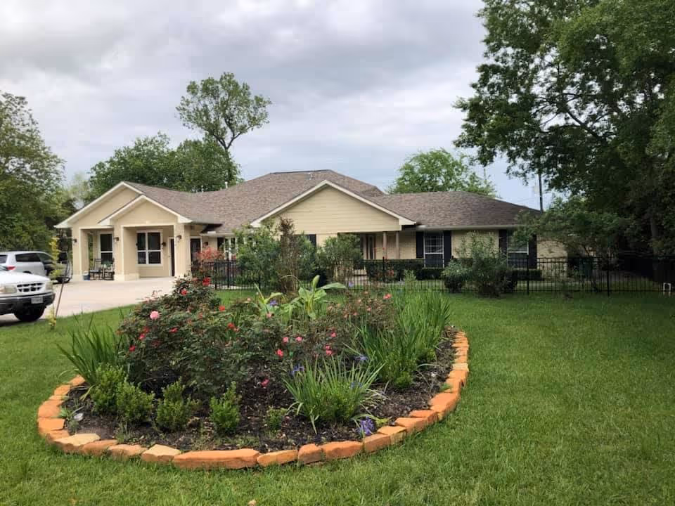 Single-story residential building with a beige exterior and a brown shingled roof, surrounded by green grass and trees. In the foreground, there is a circular flower bed bordered with bricks, containing various plants and flowers. A white vehicle is parked on the driveway to the left of the building under a cloudy sky.