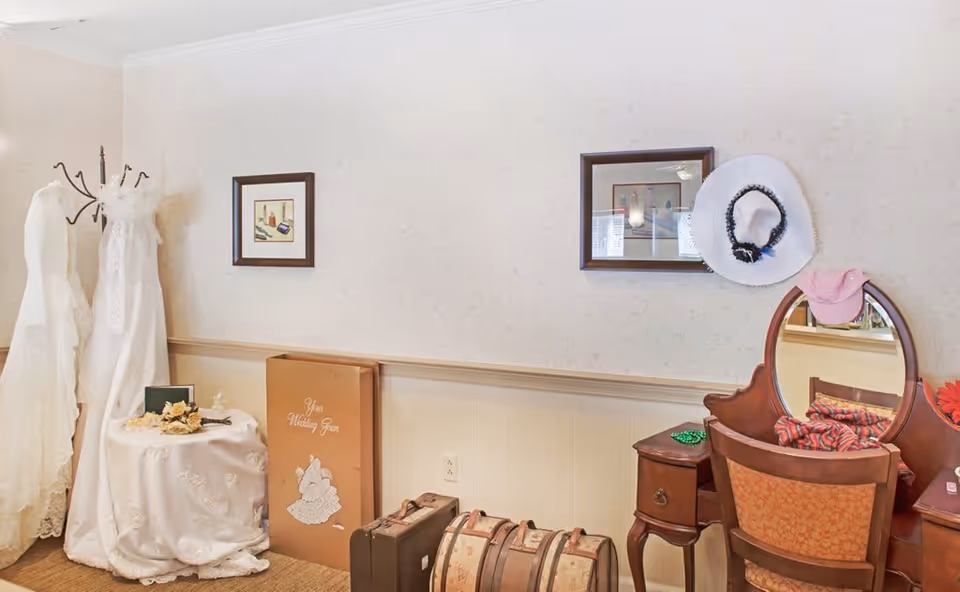 A corner of a room with two white wedding dresses on mannequins, a small round table covered with a white cloth and decorated with flowers, a large book titled 'Your Wedding Year' leaning against the wall, two vintage suitcases on the floor, and a wooden vanity with a mirror and hats hanging on it. The walls are light-colored with a subtle pattern and two framed pictures are hung above the table and vanity.