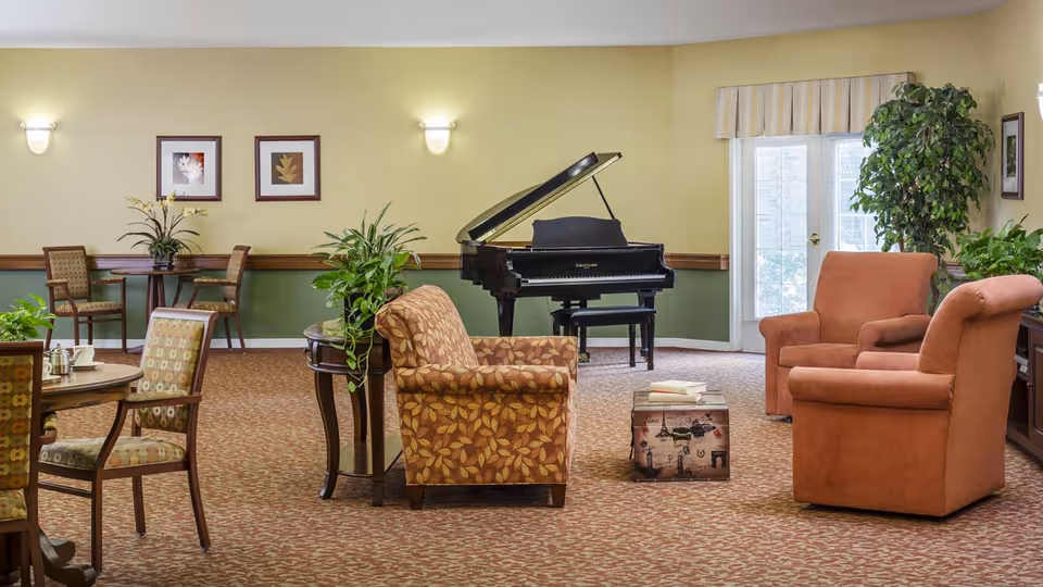 A cozy living room area with patterned carpet, two orange armchairs, a floral-patterned sofa, a small table with books, a grand piano in the background, several potted plants, framed artwork on the walls, and a glass door letting in natural light.