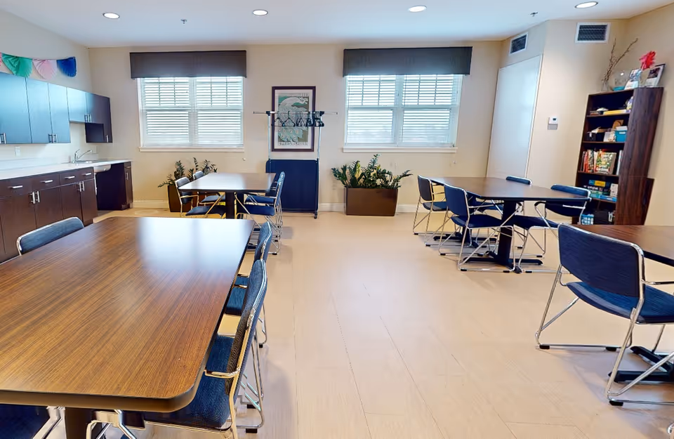 A bright, spacious room with multiple tables and chairs arranged for group seating. The room features large windows with blinds, dark wood cabinets with a sink on the left wall, and a bookshelf filled with games and books on the right. There are two large potted plants near the windows and a coat rack with hangers in the center of the back wall. The floor is light-colored, and the ceiling has recessed lighting.
