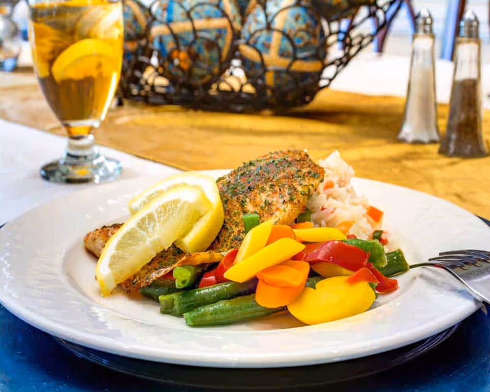 A plate of seasoned grilled fish topped with two lemon wedges, served with mixed steamed vegetables including green beans, carrots, and red and yellow bell peppers, alongside a portion of rice. In the background, there is a glass of iced tea with lemon and a decorative basket on a yellow tablecloth, with salt and pepper shakers nearby.