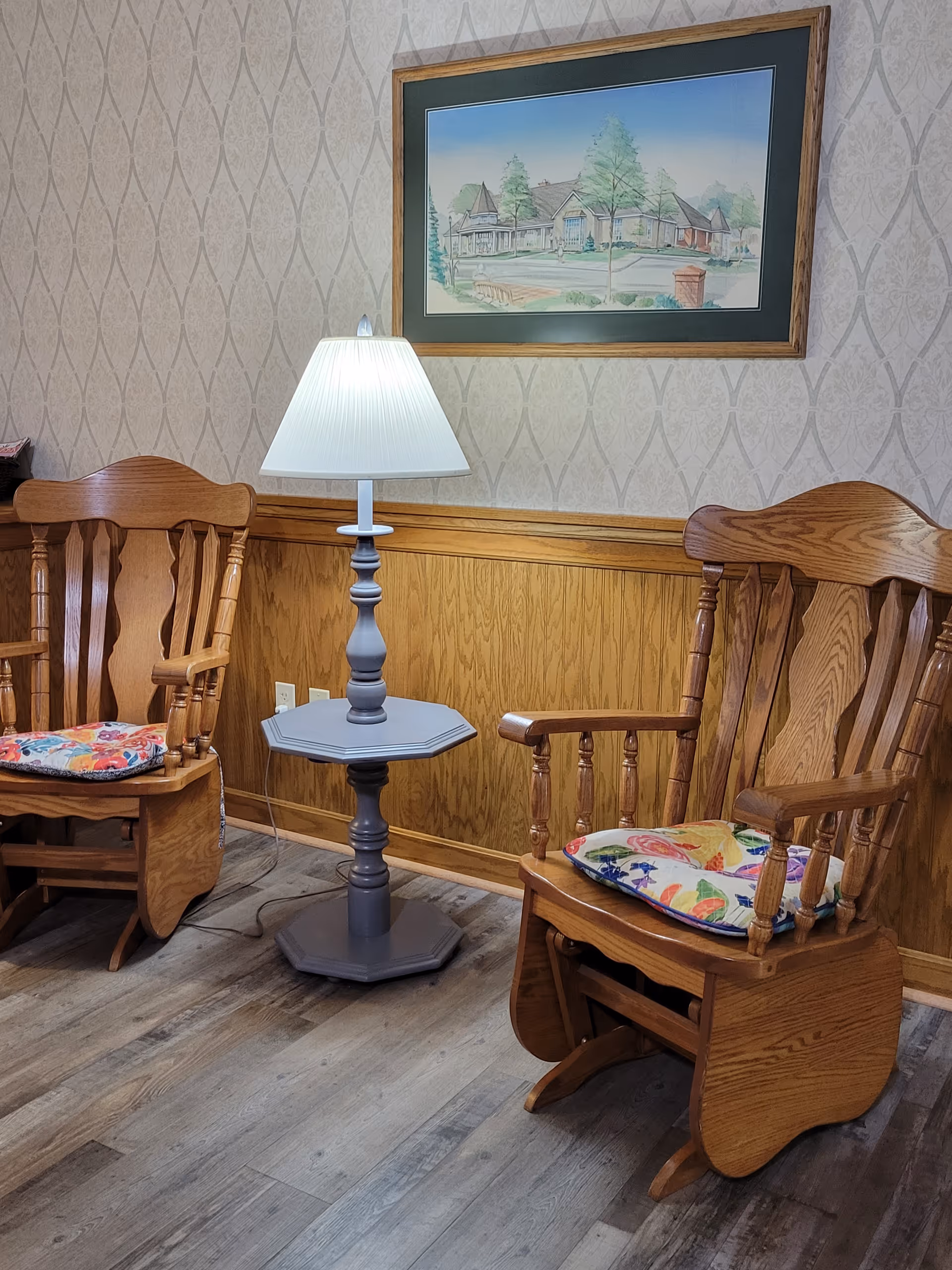 Two wooden rocking chairs with colorful cushions are positioned on either side of a small gray side table with a white lampshade. The room has wood paneling on the lower half of the wall and patterned wallpaper above. A framed painting of a house and trees hangs on the wall above the lamp.