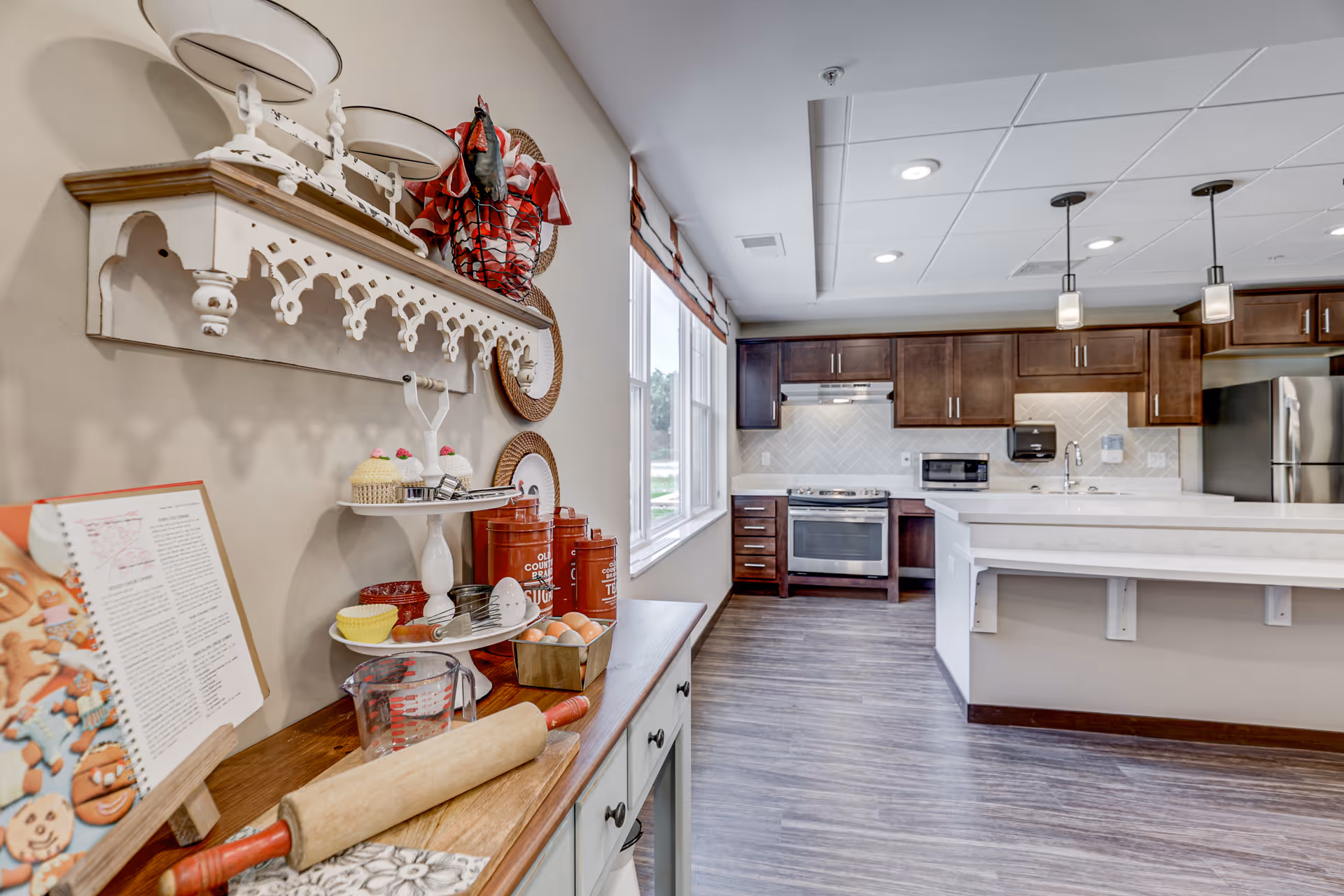 A bright kitchen area with dark wooden cabinets, a stainless steel oven and refrigerator, and a white countertop island with pendant lights hanging above. On the left side, there is a wooden sideboard with baking items including a rolling pin, measuring cup, eggs, cupcake decorations, and a cookbook on a stand. The wall above the sideboard has a decorative white shelf with plates and a red rooster figurine.