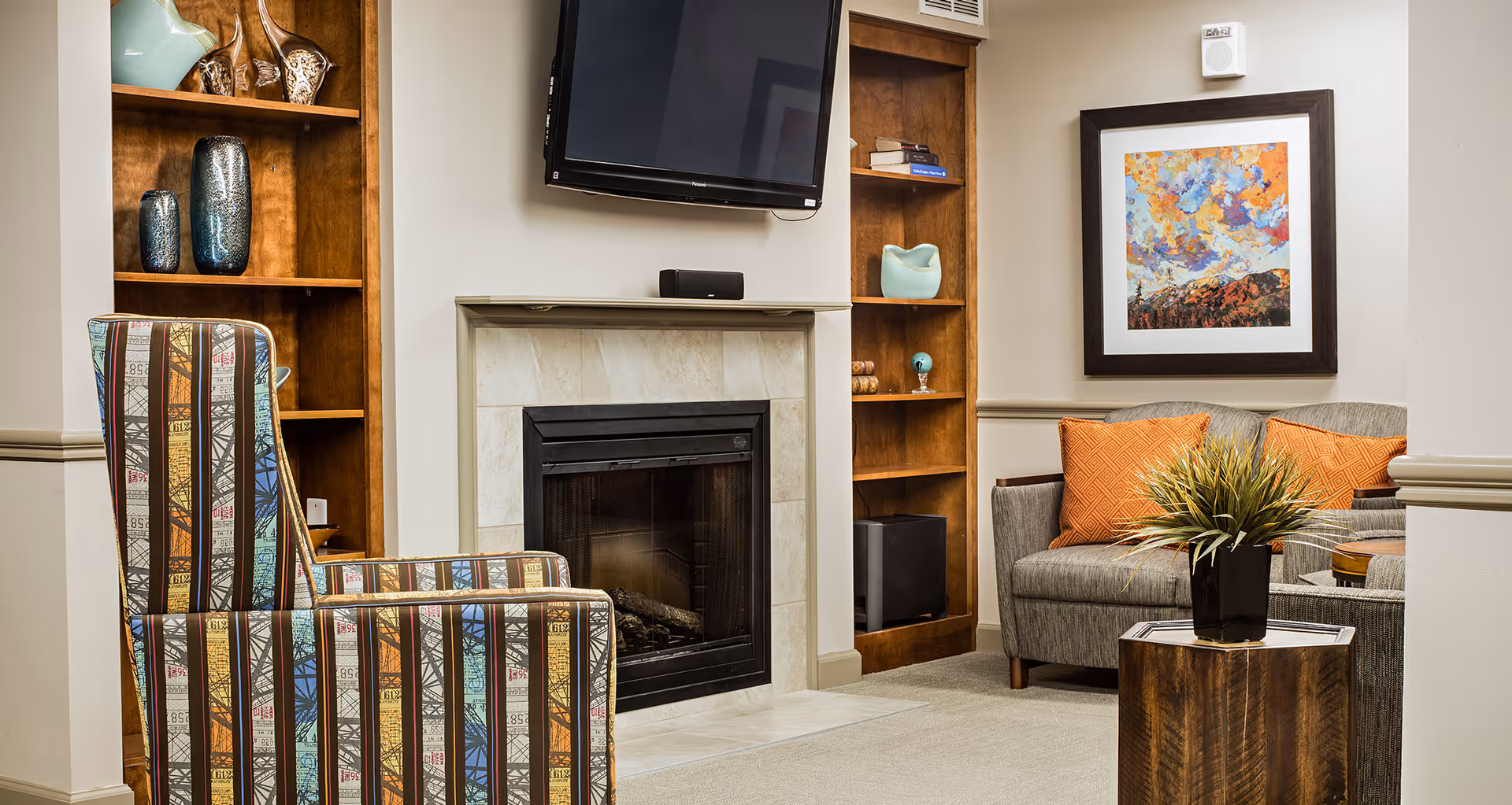 A cozy living room with a fireplace and mounted TV, patterned armchair, gray sofa with orange pillows, built-in wooden shelving, and a small wooden side table with a plant.